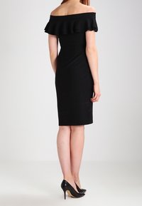 Czarna sukienka z odkrytymi ramionami, z falbanką przy dekolcie, dopasowanym krojem, o długości do kolan i gładkiej fakturze. Noszona z czarnymi szpilkami.
