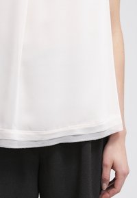 Top bianco e trasparente con taglio dritto, caratterizzato da una trama liscia e un orlo a strati. Abbinato a pantaloni drappeggiati scuri.