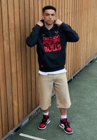 Felpa nera con logo rosso "Chicago Bulls", pantaloni corti beige, sneakers neri e rossi, in piedi contro uno sfondo di legno.
