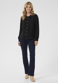 Blusa nera con un fronte arricciato, dotata di bottoni in perla, abbinata a jeans svasati scuri. Il modello si trova su uno sfondo grigio semplice.