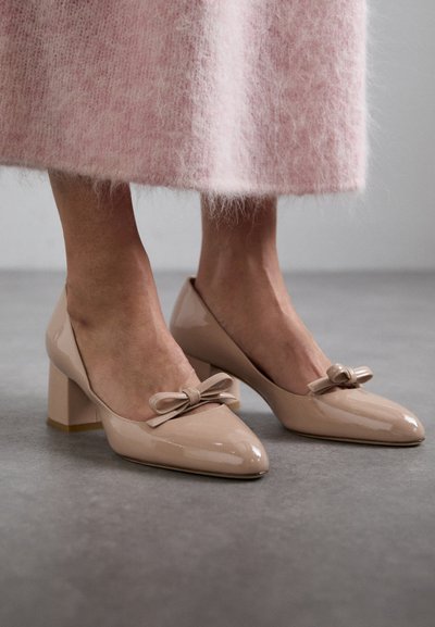 Beige Lacklederabsätze mit spitzer Zehe, dicker Ferse und einer Schleifendekoration vorne. Teilweise sichtbarer strukturierter rosa Rock.