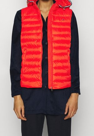 Mann trägt eine leuchtend rote, gesteppte ärmellose Steppweste über einem schwarzen Hemd mit Knöpfen und schwarzen Hosen, steht mit entspannten Armen an den Seiten.