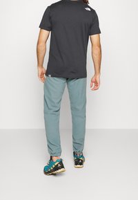 Hombre vestido con camiseta negra de The North Face, pantalones de chándal verde grisáceo y zapatillas de trail running azules de Salomon, de pie frente a un fondo liso.