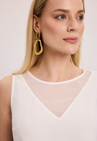 Biała bluzka z przezroczystym, w kształcie litery V dekoltem. Modelka nosi duże, żółte geometryczne kolczyki z przezroczystym detalem. Gładka faktura.