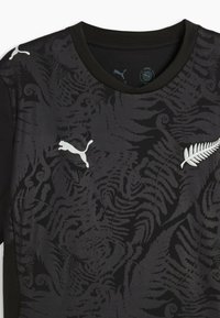 Tricou sport negru cu model de ferigă întunecată, având logo-ul alb Puma pe pieptul drept și emblema alb-argintie a ferigii pe pieptul stâng.