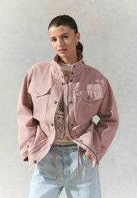 Veste en coton rose avec des boutons-pression, des poches poitrine doubles, un ourlet avec cordon de serrage et un col montant, portée sur un haut à motifs et un jean clair.
