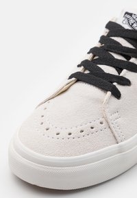 Nahaufnahme der Zehenpartie eines beigen Wildledersneakers mit Perforierungen, schwarzen Schnürsenkeln und weißer Gummisohle auf einem weißen Hintergrund.