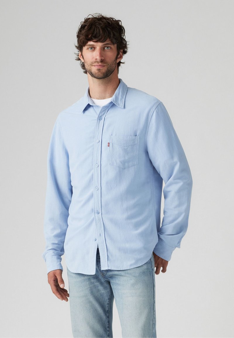 Lichtblauw overhemd met lange mouwen en knopen, gemaakt van zachte stof, voorzien van een borstzak en een iets afgeronde zoom. Gekleed met lichte jeans.