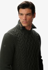Ciemnozielony sweter z dzianiny kablowej z prążkowanym dekoltem. Pod spodem widoczny jest kołnierzyk w kratę, dodający faktury i kontrastu.