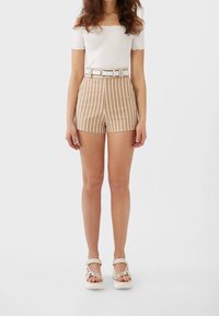 Pantaloni corti a righe beige e bianchi con vita alta, passanti per la cintura e una cintura bianca. Abbinati a un top aderente bianco scollato sulle spalle e sandali chiari.