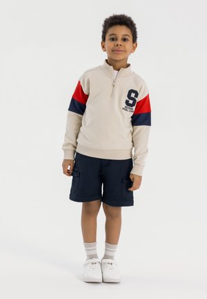 Lichtbeige ritsvest met rode en marineblauwe gekleurde mouwen, marineblauwe shorts, witte sneakers en gestreepte sokken. Voorzien van logo-detail.
