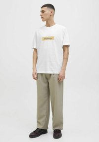 Hvid bomulds t-shirt med et gult grafisk logo. Sammen med løse, beige bukser og mørkebrune sko. Modellen står i profil.