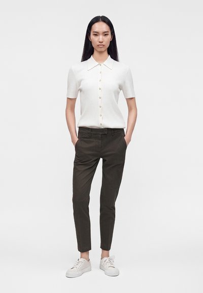Femme aux longs cheveux foncés portant une chemise blanche à manches courtes boutonnée, un pantalon slim foncé et des baskets blanches, debout les mains dans les poches.