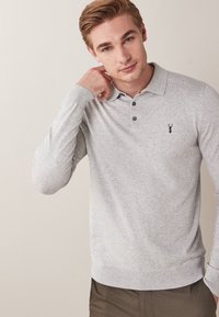 Harmaa pitkähihaisen poolopaidan, jossa on kaulus, resorit hihansuissa ja rintataskun logo. Pehmeä koostumus, edessä napit.