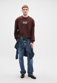 Καφέ φούτερ με λογότυπο Calvin Klein, μπλε καρό πουκάμισο δεμένο στη μέση, ανοιχτό μπλε τζιν παντελόνι και σκούρες καφέ ankle boots.