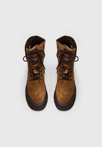 Botas de tobillo de ante marrón con una robusta suela de goma negra, diseño con cordones y un forro interior texturizado. Presenta ojales de metal y detalles de costura.