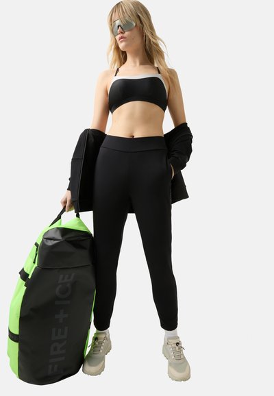 Zwarte sport-bh en hoog-zittende legging, gecombineerd met een zwarte jas. Houdt een grote, groene en zwarte sporttas vast. Grijze sneakers maken de outfit compleet.