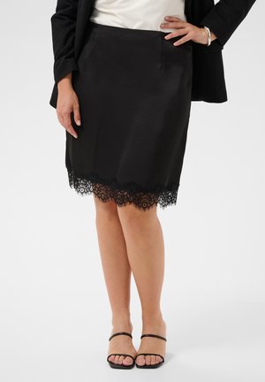 Femme portant une jupe noire à ourlet en dentelle, un haut blanc, un blazer noir, un bracelet argenté et des sandales à talons ouvertes noires, debout sur un fond blanc.