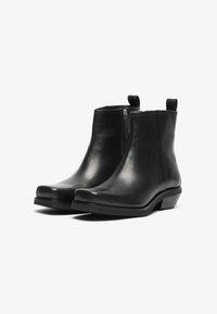 Botines de cuero negro con textura suave, cierre de cremallera y lengüetas. Presentan un tacón bajo y una punta redondeada.
