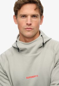 Sudadera gris claro con cuello alto, detalles con cordón y logo rojo "SUPERDRY". Tela suave y textura lisa.