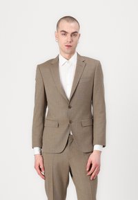Completo marrone a scacchi con design aderente, revers a punta e due tasche anteriori. Abbinato a una camicia bianca con bottoni e dettagli argentati.
