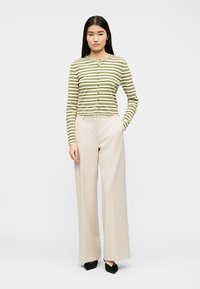 Γυναίκα που στέκεται με το χέρι στην τσέπη μπεζ παντελονιού με φαρδιά μπατζάκια, φορώντας πράσινη και λευκή ριγέ μπλούζα με κουμπιά και μακρυμάνικη, καθώς και μαύρα μυτερά παπούτσια.