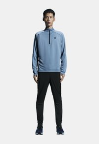 Ljust blå långärmad träningströja med kvartslång dragkedja, matchad med svarta byxor och mörka sneakers. Har en diskret logotyp på bröstet.