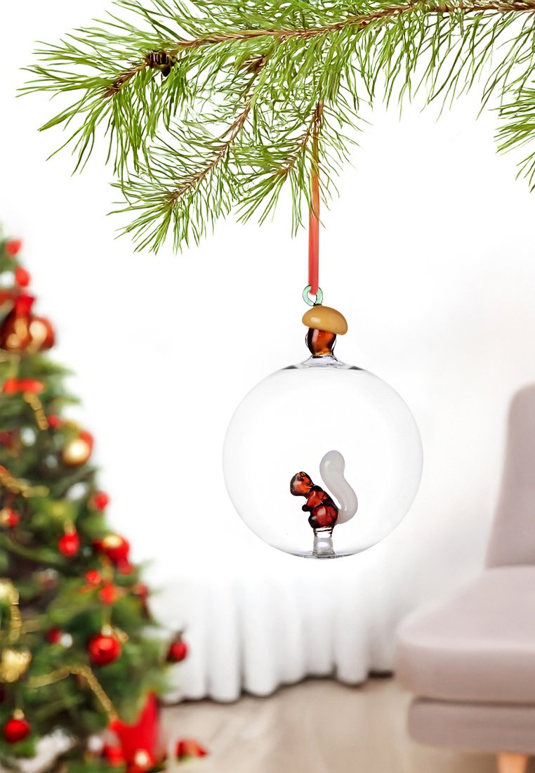 Ornamento di vetro trasparente che presenta un scoiattolo marrone con una coda bianca all'interno, decorato con una ghianda in cima e appeso a un nastro rosso tra i rami di pino.