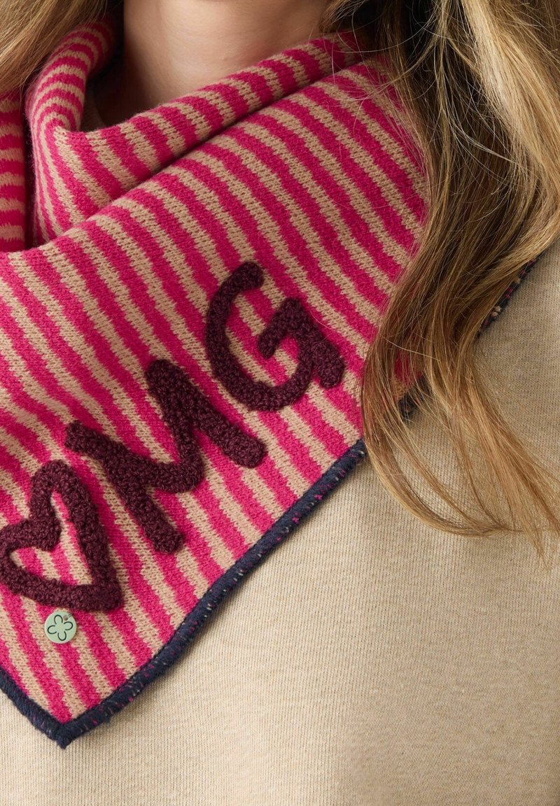 Gros plan d'une écharpe rayée rose et beige avec des lettres sombres texturées "MG" et un cœur, portée sur un pull beige et de longs cheveux châtain clair.