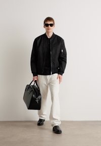 Schwarze Bomberjacke, schwarzes Hemd, weiße Hose, schwarze Sonnenbrille. Hält eine schwarze Tasche. Glattes Material, strukturierter Schnitt, grundlegende Farben.