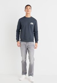 Sudadera gris oscuro con cuello redondo y puños de canalé, combinada con jeans gris claro y zapatillas blancas. Diseño simple y sin adornos.