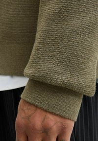 Pateešana tetovēta roka no olīvzaļā adītā džempera piedurknes, ar nelielu skatu uz svītrainām biksēm un baltu kreklu zem tā.