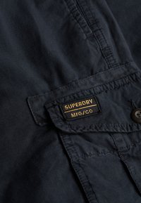 Tissu foncé avec une étiquette noire et jaune Superdry MFG/CO cousue sur un rabat de poche à boutons.