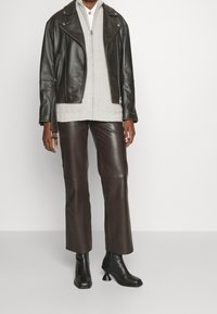 Schwarze Lederbikerjacke über einem hellgrauen gerippten Pullover, kombiniert mit dunkelbraunen weiten Lederhosen und schwarzen Absatzstiefeln mit Knöchelschnitt.