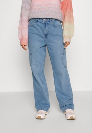 Osoba ubrana w luźne niebieskie jeansy typu cargo, wielokolorowy pastelowy sweter i białe trampki, stojąca na tle jednolitego tła.