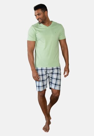 Man loopt blootsvoets en draagt een lichtgroen T-shirt met V-hals en blauw-zwart-witte geruite korte broek tegen een effen achtergrond.