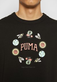 T-shirt noir avec le texte "PUMA" entouré d’abeilles, de champignons, de soleils avec le logo Puma, et des illustrations de la Terre en motif circulaire.