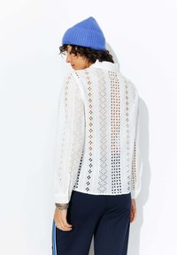 Blouse blanche à manches longues en tissu à œillets, avec un col et des poignets à boutons. Associée à un pantalon sombre et un bonnet en tricot bleu.