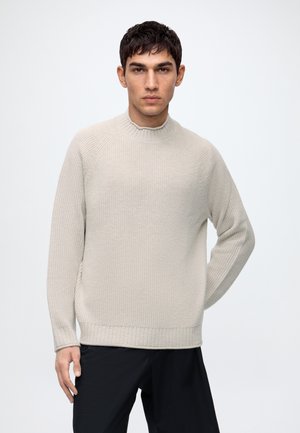 Jeune homme aux cheveux courts et foncés portant un pull en maille côtelée beige clair et un pantalon noir sur un fond clair uni.