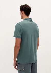 Polo skjorte i dæmpet teal stof, korte ærmer og en klassisk krave. Har en slidse i siden ved nederkanten og et lille mærke på den nederste side.