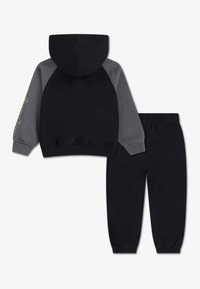 Hanorac negru cu mâneci raglan gri și glugă, având un text grafic auriu. Însoțit de pantaloni de trening negri asortați, cu manșete elastice.