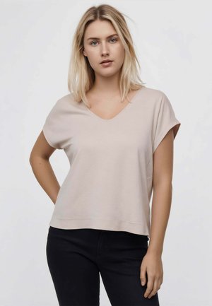 Blond kvinde med blå øjne iført en lysebeige kortærmet V-hals top og sorte bukser, står med den ene hånd på hoften.