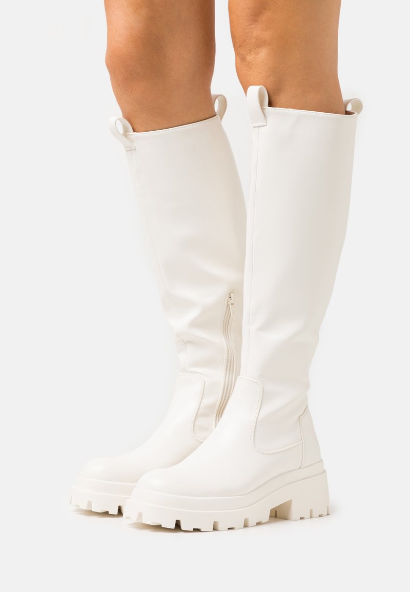 Bottes blanches montantes en matériau synthétique lisse, avec une semelle épaisse, une fermeture éclair sur le côté et des accents de tirette.