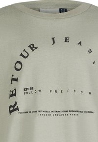 Hellgrünes Baumwoll-T-Shirt mit schwarzem Textdesign, einschließlich "RETOUR JEANS" und "FOLLOW FREEDOM", mit Rundhalsausschnitt und kurzen Ärmeln.