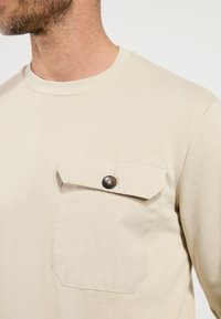 Beige langemouw overhemd van zacht materiaal, met een knoopzak op de borst met een ronde metalen drukknoopsluiting en een ronde hals.