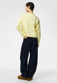 Sudadera amarilla con cuello redondo y mangas largas, combinada con pantalones vaqueros de pierna ancha de talle alto en azul oscuro y calzado marrón con plataforma.