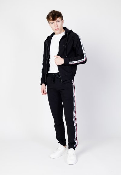 Czarna bluza z zamkiem błyskawicznym i białymi paskami tekstowymi, noszona na białej koszulce. Połączona z czarnymi dresami z pasującymi paskami po bokach i białymi sneakersami.