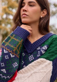 Šarena pulover od zakrpa s dubokim V-izrezom, teksturiranim pletenim uzorcima u mornarsko plavoj, zelenoj, krem i crvenoj boji s geometrijskim dizajnom.