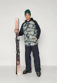 Ski apranga, sudaryta iš maskuojančio gobtuvo, kuriame dominuoja žali ir jūriniai tonai, derinama su tamsiomis kelnėmis, juodais slidinėjimo batais ir laikoma slidėmis su tvirtinimais.