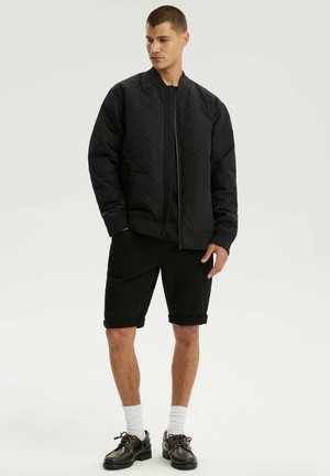 Mężczyzna w czarnej pikowanej kurtce typu bomber, czarnych podwiniętych szortach, białych skarpetkach i czarnych skórzanych butach stojący na jednolitym tle.